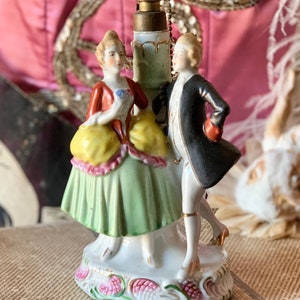 May include: A porcelain figurine of a couple dancing. The woman is wearing a green and yellow dress with a red bodice. The man is wearing a black coat and white pants. The figurine is on a white base with pink and green floral details.