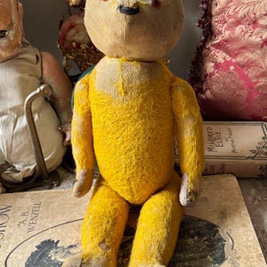 May include: A vintage yellow teddy bear with a stitched face, button eyes, and a black nose. The bear is sitting on a book with the text "DRAWINGS BY A.B. WENZELL". The bear's fur is worn, giving it a classic, antique appearance.