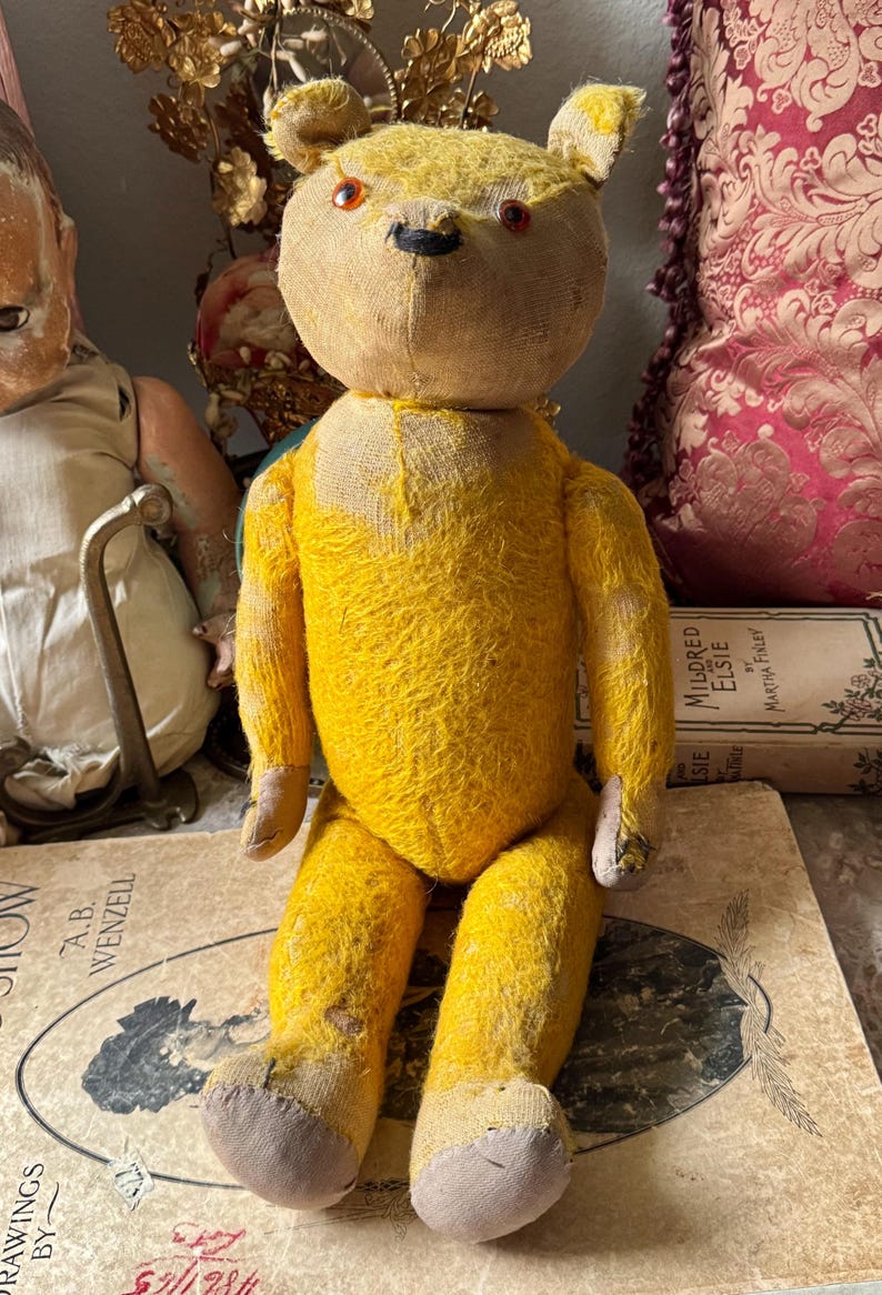 May include: A vintage yellow teddy bear with a stitched face, button eyes, and a black nose. The bear is sitting on a book with the text "DRAWINGS BY A.B. WENZELL". The bear's fur is worn, giving it a classic, antique appearance.