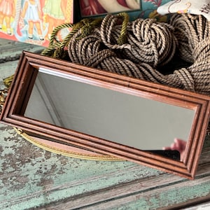 May include: A rectangular brown wooden framed mirror with a reflective surface. The mirror is resting on a decorative gold-colored tray. A bundle of rope and other decorative items are in the background.