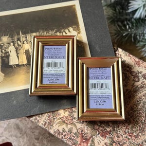 May include: Two small, rectangular gold-colored photo frames with a purple matting. Each frame has the text "PHOTO FRAME" and "INTERCRAFT" printed on the label. The frames measure 2.25 x 3.25 inches (6 x 8 cm).