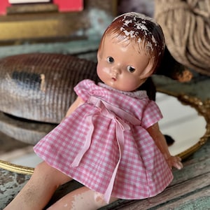 May include: A vintage doll with a painted face and brown hair, wearing a pink and white checkered dress with a bow. The doll is resting on a gold-framed mirror. A wooden duck and rope are in the background.