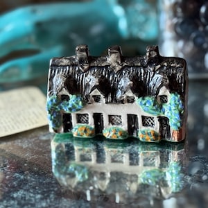 May include: A miniature ceramic cottage with a thatched roof, featuring a white facade with windows and colorful shrubbery. The cottage is reflected on a shiny surface, creating a mirrored effect. The cottage is a decorative item.