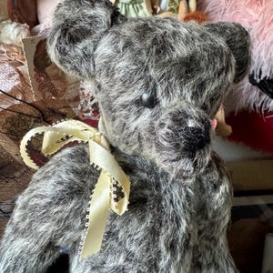 May include: A vintage-style teddy bear with gray and white fur. The bear has a dark nose and eyes, and a cream-colored ribbon tied around its neck. The bear is surrounded by other vintage items, suggesting a collectible or antique theme.
