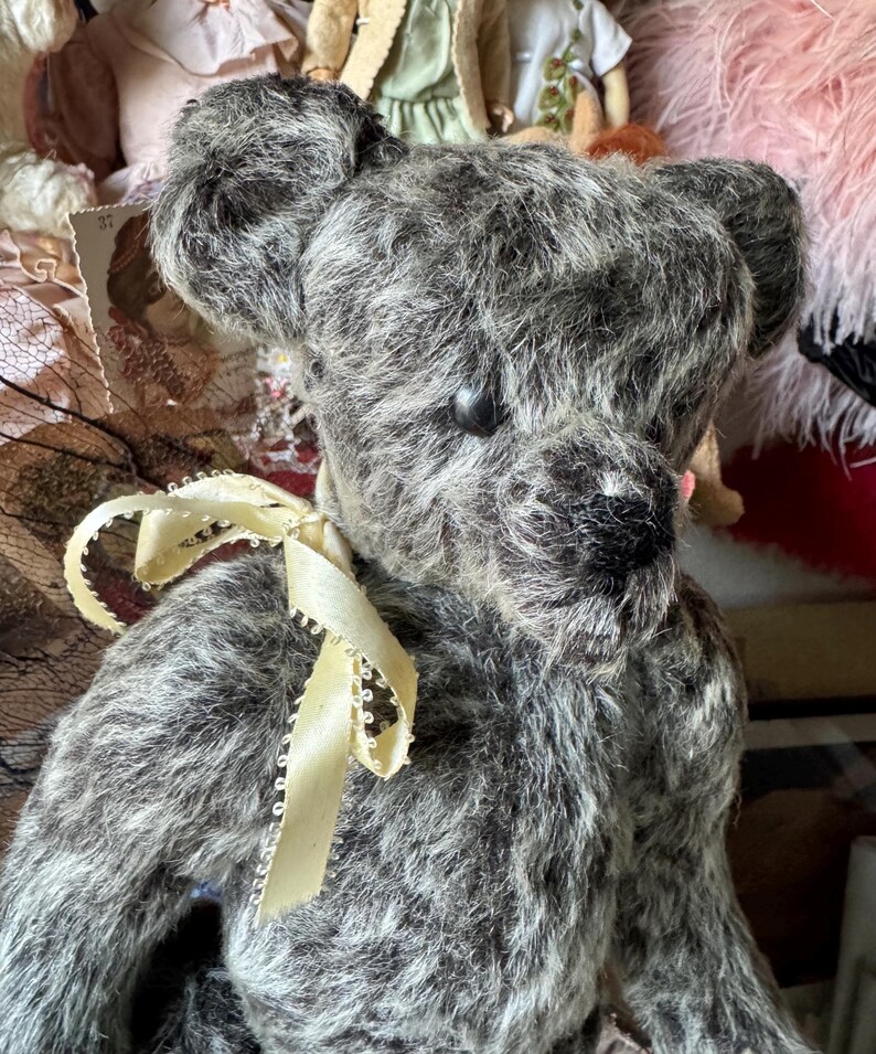 May include: A vintage-style teddy bear with gray and white fur. The bear has a dark nose and eyes, and a cream-colored ribbon tied around its neck. The bear is surrounded by other vintage items, suggesting a collectible or antique theme.