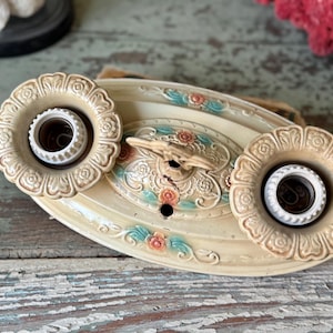 May include: Cream-colored vintage ceiling light fixture with two bulb sockets. The fixture has an oval shape with floral and scrollwork details in pink and teal. The light fixture is made of metal.