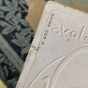 Pode incluir: Close-up de um cart&atilde;o vintage branco sujo com desenhos florais em relevo e o texto "Series 226 A" em preto. As bordas do cart&atilde;o mostram sinais de desgaste, sugerindo sua idade. O fundo apresenta um tecido estampado.