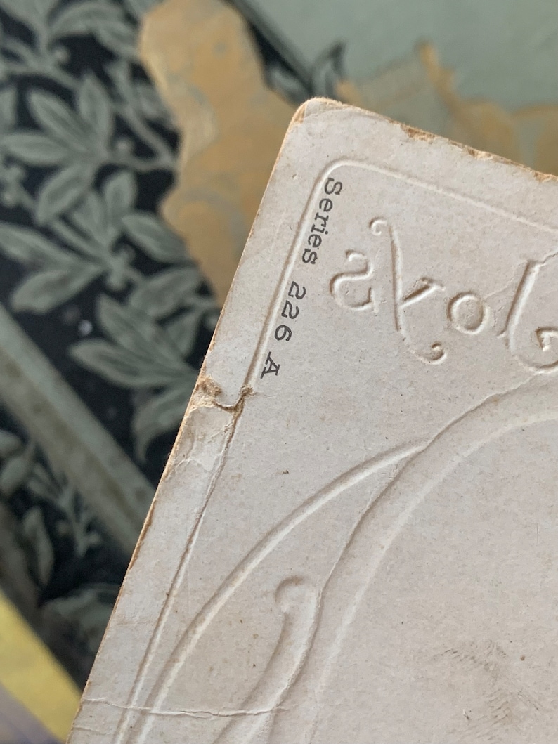 Pode incluir: Close-up de um cart&atilde;o vintage branco sujo com desenhos florais em relevo e o texto "Series 226 A" em preto. As bordas do cart&atilde;o mostram sinais de desgaste, sugerindo sua idade. O fundo apresenta um tecido estampado.