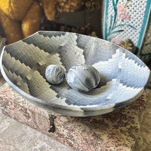 May include: A shallow, wide bowl featuring a geometric, mosaic-like pattern in shades of gray, white, and black. Inside the bowl are two smooth, rounded stones with a marbled, swirling gray and white texture that matches the bowl's color palette.