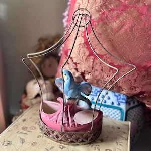 May include: A decorative birdcage-shaped planter with a blue ceramic bird perched inside. The planter has a pink base and a metal cage structure. The bird has blue and white coloring. The cage sits on a floral patterned surface.