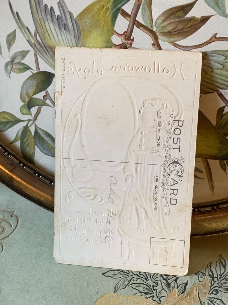 Pode incluir: Um postal vintage com detalhes em relevo. O cart&atilde;o apresenta as palavras "Halloween Greetings" e "Post Card" em uma fonte decorativa. A parte de tr&aacute;s do cart&atilde;o tem uma apar&ecirc;ncia desbotada e envelhecida, com um fundo floral.