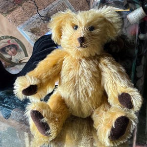 May include: A vintage-style teddy bear with golden fur and dark brown paws and nose. The plush toy has a classic design with a slightly scruffy appearance, giving it a well-loved look. The bear is sitting, with its arms and legs outstretched.