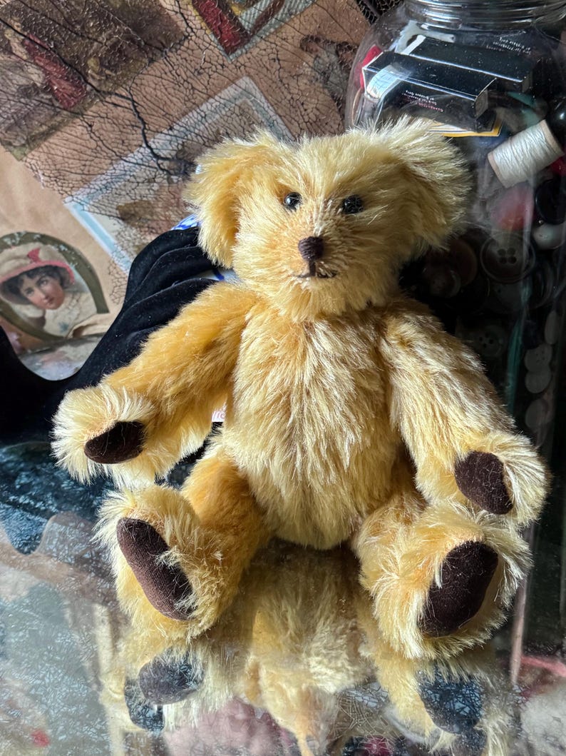 May include: A vintage-style teddy bear with golden fur and dark brown paws and nose. The plush toy has a classic design with a slightly scruffy appearance, giving it a well-loved look. The bear is sitting, with its arms and legs outstretched.