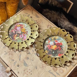 May include: Two circular wall frames featuring ornate gold-toned metal borders decorated with a repeating butterfly motif. Each frame contains a colorful floral still life print depicting a bouquet of flowers in a vase, set against a light background.