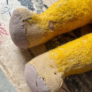 May include: Close-up of two yellow plush toy legs with gray fabric feet. The legs have a worn, aged appearance with visible patches and stitching. The background features a vintage paper with faded illustrations.