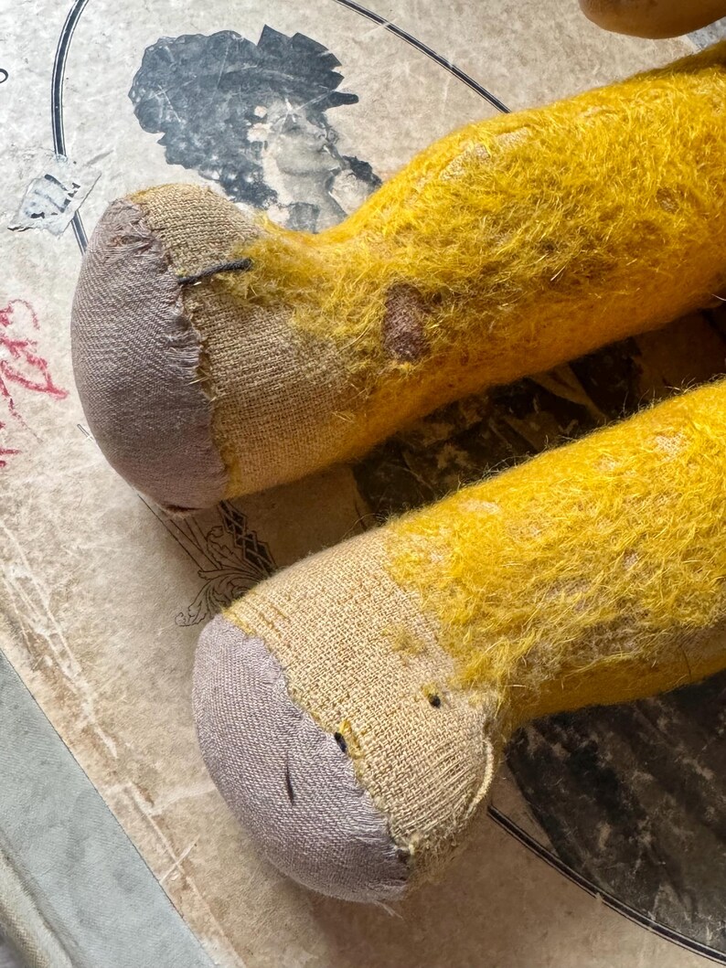 May include: Close-up of two yellow plush toy legs with gray fabric feet. The legs have a worn, aged appearance with visible patches and stitching. The background features a vintage paper with faded illustrations.