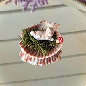 May include: A small white ceramic bunny figurine is nestled in a bed of green moss, surrounded by a stack of brown seashells. A small red and white mushroom sits next to the bunny.