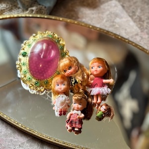 May include: A collection of miniature dolls with blonde hair and various outfits, arranged on a reflective surface. The dolls are in dresses of red, white, and plaid patterns. A decorative, ornate oval-shaped object with a pink center is also present.