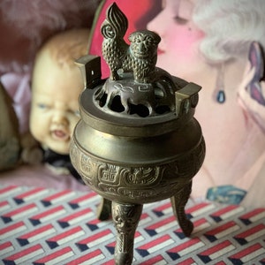 May include: A bronze incense burner with a decorative lion on top. The burner has a round body with intricate carvings and three ornate legs. The background includes a doll and a vintage-style illustration.