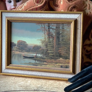May include: A framed painting of a lake scene with trees and a small boat in the water. The painting is in a simple gold and white frame.