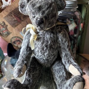 May include: A vintage-style teddy bear with gray fur and a cream-colored ribbon around its neck. The bear has a classic design with jointed limbs and a soft, plush texture. Its paws have tan pads.