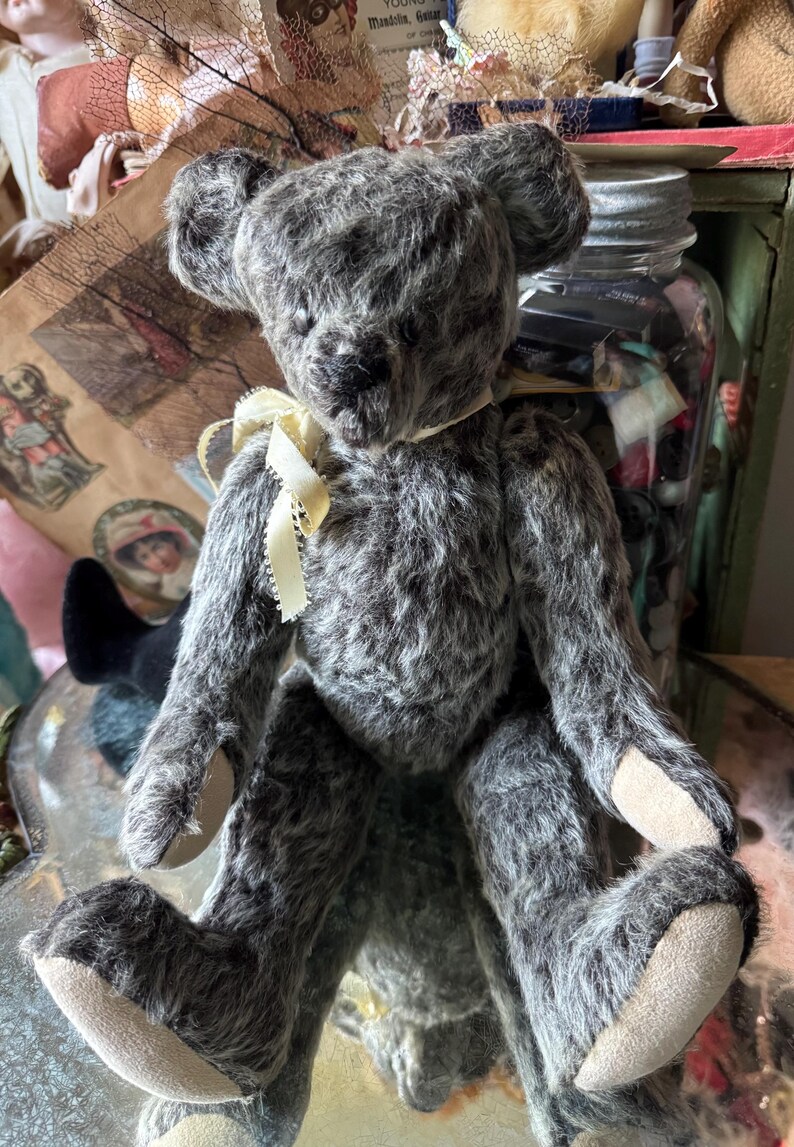 May include: A vintage-style teddy bear with gray fur and a cream-colored ribbon around its neck. The bear has a classic design with jointed limbs and a soft, plush texture. Its paws have tan pads.