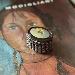 May include: An oval, antique silver-toned trinket box with a mirrored top. The box features ornate detailing around the sides and a reflective surface. The box is set on a vintage magazine with the word "MODIGLIANI" visible.