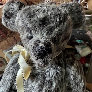 May include: A handmade gray teddy bear with a cream-colored ribbon tied around its neck. The bear has a textured, shaggy fur appearance and black button eyes. The background is blurred, suggesting a vintage or antique setting.