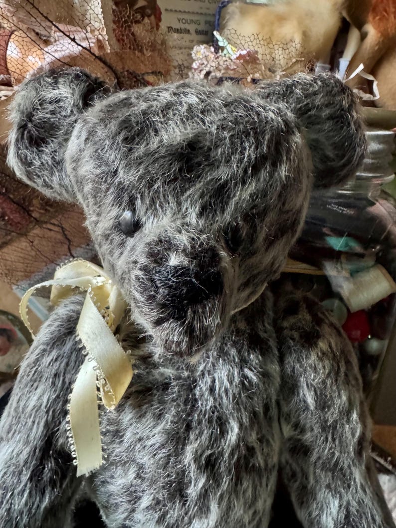 May include: A handmade gray teddy bear with a cream-colored ribbon tied around its neck. The bear has a textured, shaggy fur appearance and black button eyes. The background is blurred, suggesting a vintage or antique setting.