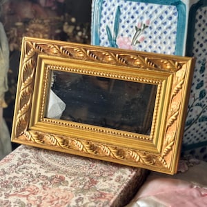 May include: A rectangular gold-colored picture frame with a mirror. The frame has an ornate, detailed design. The frame is resting on a floral patterned box. The background includes a doll and floral patterned objects.