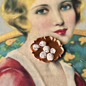 May include: A copper-colored, scalloped-edge dish holds several small, white, textured objects. The dish is set against a vintage illustration of a woman with blonde hair, blue eyes, and red lipstick.