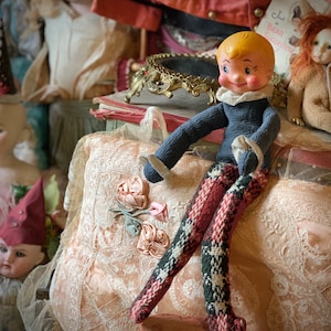 May include: A vintage doll with a blue sweater and red and white striped pants sits on a lace tablecloth. The doll has a painted face with blonde hair and blue eyes.