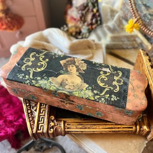 May include: A rectangular wooden box with a hinged lid featuring a central illustration of a woman in a hat, surrounded by black and gold scrollwork and green floral patterns on a peach-colored background, resting on a gold-toned stand.