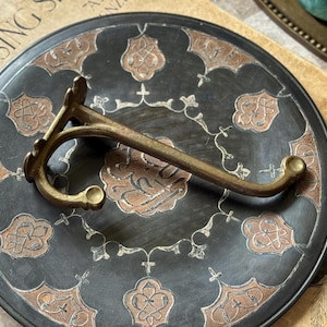 May include: An antique brass wall hook with a curved design, resting on a decorative black plate with copper-colored floral patterns. The hook has a vintage aesthetic, suggesting a focus on home decor and organization.