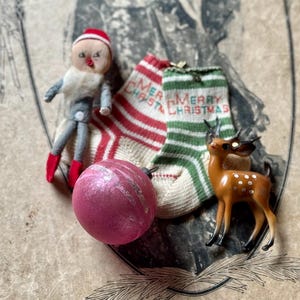 May include: A collection of vintage Christmas decorations. Includes a small doll with a red hat and boots, two miniature Christmas stockings with "Merry Christmas" text, a pink ornament, and a small deer figurine.