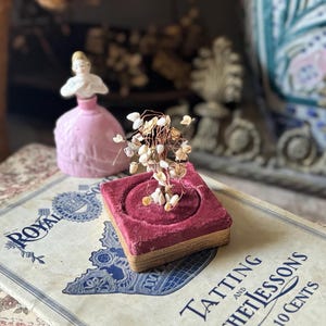 May include: A small wire tree sculpture with white shell accents sits on a square, deep red velvet pin cushion with gold trim. Beside it is a pink porcelain figurine of a person in a dress, placed on a vintage book titled Royal Tatting and Crochet Lessons.