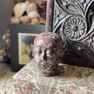 May include: A weathered, antique-style sculpture of a human head with closed eyes. The head is painted in shades of red and white, with visible texture. The sculpture is placed on a floral patterned surface, with a dark wooden carved panel in the background.
