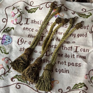 May include: Four olive green decorative tassels with twisted cords and knotted tops. The tassels are lying on a cream-colored embroidered fabric with floral and text details. The text reads "I shall as soon..."