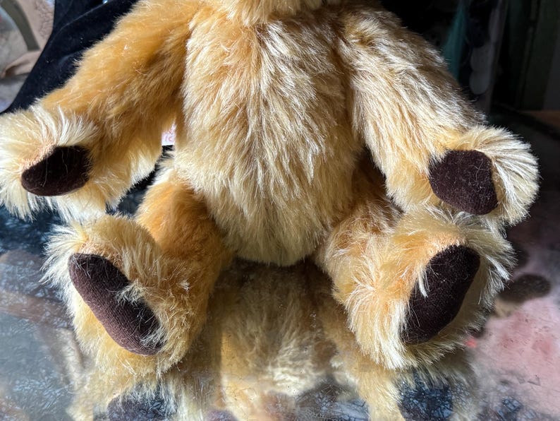 May include: A plush, golden-brown teddy bear with dark brown paw pads. The bear has a soft, fluffy texture and is sitting with its arms and legs extended. The fur is a mix of light and medium brown tones.