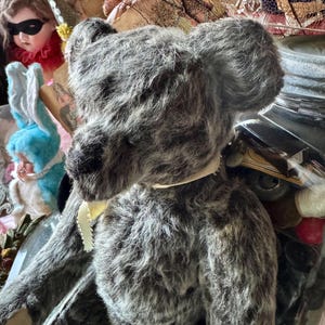 May include: A vintage-style gray teddy bear with a cream-colored ribbon around its neck. The bear has a textured, plush appearance and dark eyes. Other antique dolls and decorative items are visible in the background.