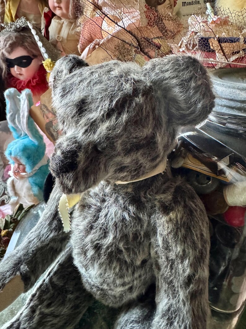 May include: A vintage-style gray teddy bear with a cream-colored ribbon around its neck. The bear has a textured, plush appearance and dark eyes. Other antique dolls and decorative items are visible in the background.