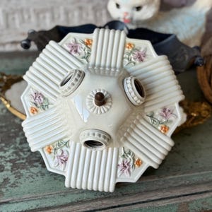 May include: Off-white, vintage ceiling light fixture with three bulb sockets. The hexagonal fixture has a ribbed design with floral accents in pink and orange. A central, decorative screw is visible.