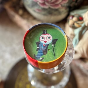 May include: A vintage toy with a colorful, circular design. The top features a cartoon figure in a suit and top hat on a green background. The rim is painted in red, yellow, and blue. The toy is set on a clear glass base.