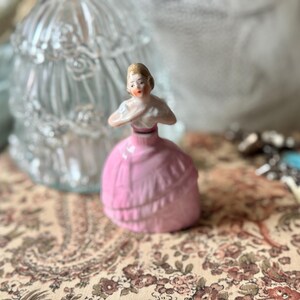 May include: A small, vintage porcelain figurine of a woman in a pink dress. The figurine has a detailed face and is posed with her arms crossed. The dress has gold accents. A clear glass container is in the background.