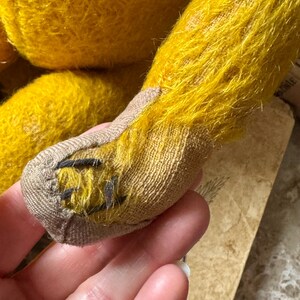 May include: Close-up of a vintage teddy bear's leg. The bear's fur is a vibrant yellow, with a textured appearance. The foot is made of a tan fabric, with black stitching details. The bear's leg is held in a hand.