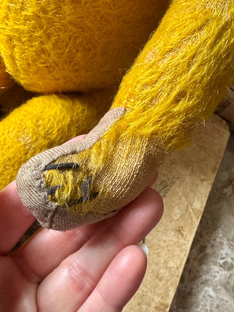 May include: Close-up of a vintage teddy bear's leg. The bear's fur is a vibrant yellow, with a textured appearance. The foot is made of a tan fabric, with black stitching details. The bear's leg is held in a hand.