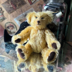 May include: A small, golden-yellow teddy bear with brown paws and a matching nose. The bear is sitting on a reflective surface, with a blurred background featuring vintage ephemera and a glass jar filled with sewing supplies.