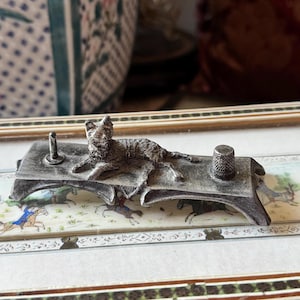 May include: A silver-toned metal cat-shaped sewing tool rest. The cat is lying down on a small table with a thimble and a pin holder. The table is set on a decorative frame with a painted scene.