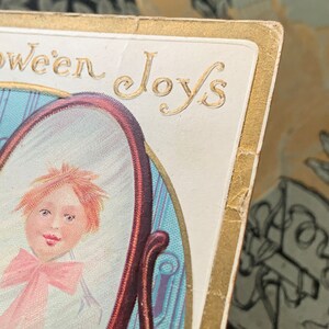 Pode incluir: Postal vintage de Halloween com o retrato de uma crian&ccedil;a num espelho. O cart&atilde;o tem bordas douradas e as palavras "Halloween Joys" em letras douradas. A crian&ccedil;a tem cabelo ruivo e um la&ccedil;o rosa.