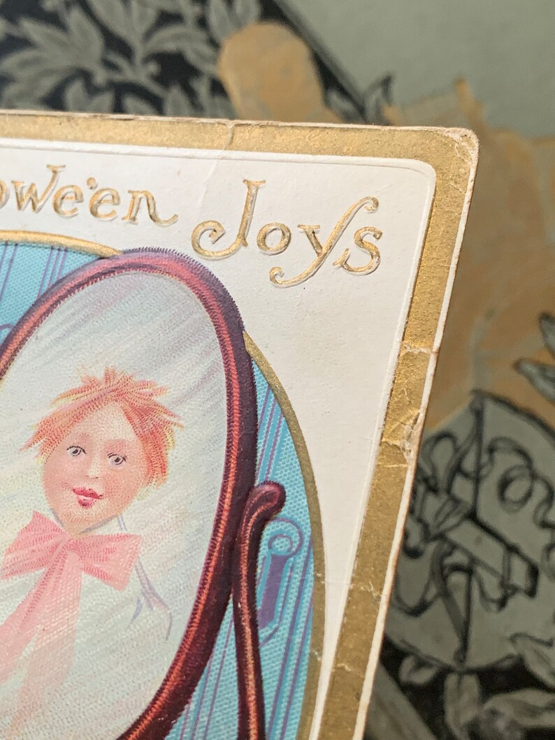 Pode incluir: Postal vintage de Halloween com o retrato de uma crian&ccedil;a num espelho. O cart&atilde;o tem bordas douradas e as palavras "Halloween Joys" em letras douradas. A crian&ccedil;a tem cabelo ruivo e um la&ccedil;o rosa.
